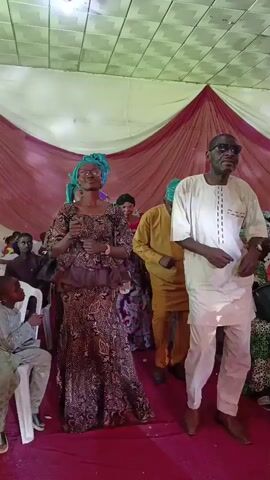 A woman in a brown and gold patterned dress and teal headwrap and a man in a white tunic and pants dance in a brightly lit tent with a red carpet. Both wear sunglasses. Other people are behind them in similar attire, and children sit in chairs to the left. Maroon and white draping decorates the walls.