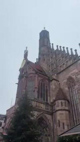 The video shows a low-angle, static shot of the side of a large, ornate, medieval church. The building is constructed of reddish-brown stone and features a clock tower topped with a golden spire. Gothic architectural elements are evident, including pointed arches, ribbed vaults, and decorative spires. In front of the church is a Christmas tree, and other trees can be seen nearby. The sky is overcast and grey, indicating a cloudy day.
