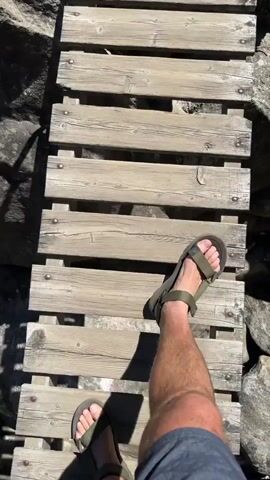 The video begins with a high-angle shot looking down at a person's feet and legs as they walk across a narrow wooden bridge. The bridge is made of weathered wooden planks laid across some sort of support structure. The person is wearing dark colored shorts and olive green sandals. On either side of the bridge are large rocks. The camera angle shifts slightly as the person continues to walk, showing more of the rocks and the bridge. The lighting suggests it is daytime, with sunlight casting shadows. The video ends with the person's feet still on the bridge, with more wooden planks visible ahead.