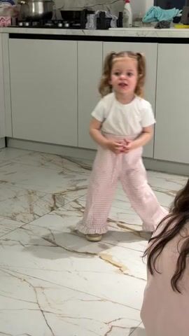 A young girl wearing a white shirt, pink and white checkered pants, and pigtails stands in a kitchen with marble floors. She is looking at the camera and makes a few gestures with her hands. A girl with long brown hair is crouched down in front of her. The kitchen has white cabinets and a variety of items on the counter, including a pot, bottles, and a cloth. Music is playing in the background.