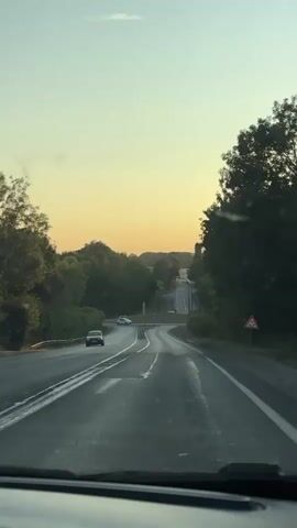 The video shows a view from inside a vehicle traveling on a road during what appears to be either sunrise or sunset. The sky is a gradient of pale yellow and light blue, with a single small cloud visible. The road is paved and appears to be wet, with white lane markings. Trees and foliage line both sides of the road, creating a tunnel-like effect. In the distance, the road curves and goes uphill, with a few other vehicles visible. A roundabout with traffic signs is also visible. The overall impression is of a serene, natural landscape with man-made infrastructure.