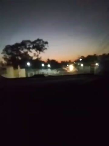 The video shows a view from inside a vehicle at dusk. The sky is dark with a hint of orange on the horizon. Trees and streetlights are visible in the distance, blurred due to the motion of the vehicle. The interior of the car is dark, obscuring the occupants. The scene appears to be a road at night with some ambient light from street lamps and possibly approaching headlights.