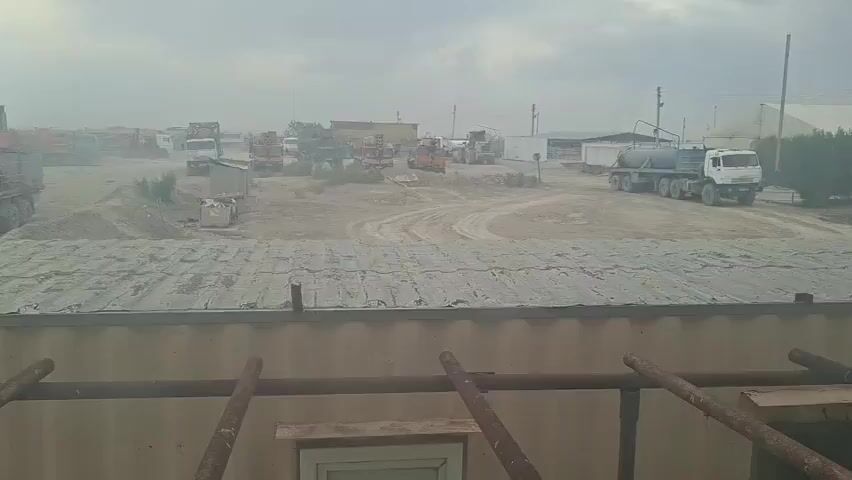 A dusty, outdoor scene of a construction or industrial yard under a hazy, overcast sky. In the foreground, a portion of a corrugated metal roof is visible, supported by metal scaffolding. The middle ground features a dirt lot with tire tracks, scattered piles of dirt, and various pieces of construction equipment and trucks parked or in motion. Several trucks are lined up on the left, some appear to be dump trucks. A water truck with a large tank is prominent on the right side of the lot. In the background, more structures and vehicles are visible, as well as some vegetation. The overall impression is one of an active, albeit dusty, work environment.