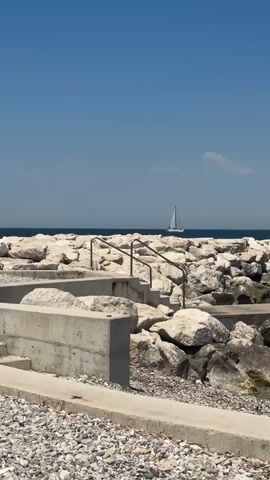 A vertical, static shot shows a rocky shoreline on a clear, sunny day. The water is blue and calm, with a sailboat visible in the distance on the horizon. In the foreground, there are concrete structures and large rocks forming a breakwater. Stairs lead down from a concrete path to the water's edge among the rocks. The sky is a bright, cloudless blue.