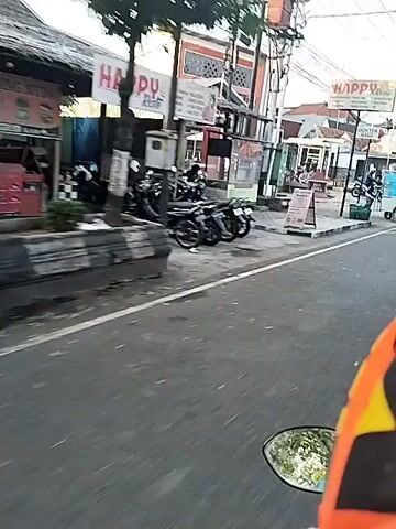 The video shows a street scene with several motorcycles and people. In the foreground, a motorcycle is parked. To the left, a person on a motorcycle is seen. Further down the street, another motorcycle is parked. A person is seen pushing a cart with wheels. In the background, there are buildings and trees. The sun is shining brightly, casting a glare on the scene. There are also some signs visible, one of which appears to be for a doctor's office. The road is paved and has white lines marking the lanes.