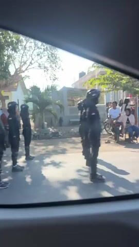 The video shows an outdoor scene through the windows of a vehicle. In the foreground, a person wearing a black uniform with a green backpack walks away from the camera. Other individuals in similar black uniforms are also visible, standing in a line on the right side of the road. On the left side of the road, a group of people are gathered, some appearing to be wearing headscarves. Further down the street, a person is seen riding on the back of a motorcycle, and other motorcycles are parked. In the background, houses and trees line the street, under a bright sky. The camera pans and shifts focus, offering glimpses of the environment and the people within it, with some individuals appearing to be in a public gathering or event.