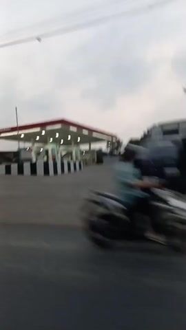 A slightly blurry video shows a gas station from the perspective of a moving vehicle. The gas station has a red and white canopy over the pumps. There are several cars and motorcycles in the lot. The sky is cloudy. In the foreground, a motorcycle passes by, also blurred due to motion. A sign for "PERTAMINA" is visible.