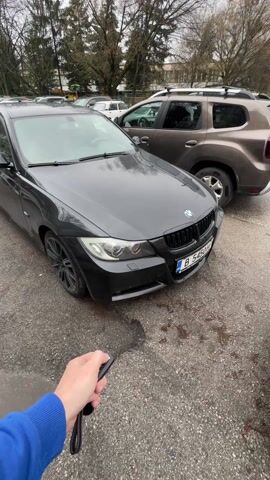The video shows a man pressing a car key, and then a quick scan of the parking lot showing a variety of cars. The first car shown is a black BMW, followed by a blue Lancia with copper-colored accents. The camera pans across an asphalt parking lot, filled with a dense row of cars, including a silver Volkswagen, and a mix of sedans and hatchbacks in various colors. The setting appears to be an urban residential area, with buildings visible in the background, suggesting a typical city parking arrangement. The weather seems overcast.