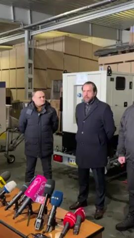Three men stand in a warehouse. In front of them is a table with an array of microphones. Behind them is a white generator on a trailer. Behind that are rows of boxes. The man in the middle wears a dark coat, gray pants, and brown shoes. The man to his left wears a dark blue puffer jacket with a brown neck, and dark pants. The man on the right wears a dark jacket with a hood, and dark pants. The man in the middle has his hands behind his back. The man on the left looks sternly to the left. The man on the right is mostly off camera.