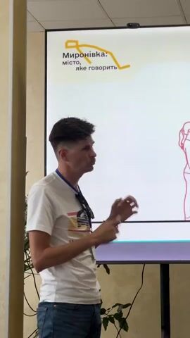 A man is standing in front of a large screen, gesturing with his hands. He is wearing a white t-shirt with a graphic design and blue jeans. He has dark hair and appears to be speaking. A lanyard with a badge is around his neck, and sunglasses are hanging on his shirt. The screen displays text in Ukrainian, with the words "Миронівка: місто, яке говорить" (Myronivka: the city that speaks). Below this, there is a graphic that appears to be a map or a path. At the bottom of the screen, there is a navigation element showing "18/23 >". The setting appears to be an indoor presentation or conference room, with a wall behind the man and a plant in the foreground. The lighting suggests it is daytime.