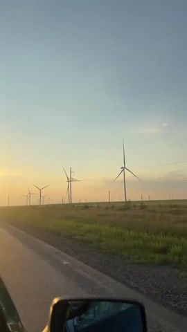 The video shows a roadside view of a wind farm at sunset. In the foreground, the side mirror of a vehicle is visible, reflecting the sky and some of the landscape. The road runs parallel to a field of tall, dry grass. A line of wind turbines with their blades rotating is visible in the distance, stretching across the horizon. Electrical pylons are also present, interspersed with the turbines. The sky is a gradient of pale blue at the top, transitioning to a soft yellow and orange near the horizon, indicating either sunrise or sunset. The lighting suggests it is either early morning or late evening. No people are visible in the frame.