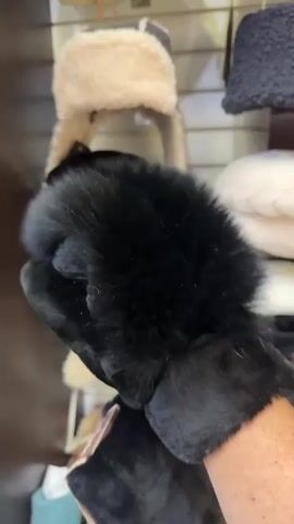 A hand wearing a black fur-lined glove is holding up a black fur-lined mitten. In the background, various hats are displayed on shelves.