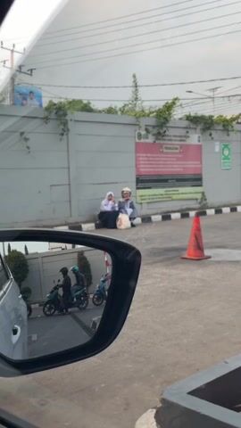 The video shows a street scene from the perspective of a car. In the foreground, the side mirror of the car reflects three people on motorcycles. One motorcycle has two riders, and the other has one rider. In the background, two people, a woman and an older man, are sitting on the ground next to a grey wall. The woman is wearing a white hijab, and the man is wearing a white head covering and a patterned shirt. They are both looking in the same direction. An orange traffic cone is placed on the pavement in front of them. Behind the two sitting people is a large sign attached to the wall, with red and white lettering. The environment appears to be urban, with a cloudy sky and some greenery growing on the wall. The overall impression is a snapshot of everyday life on a street.