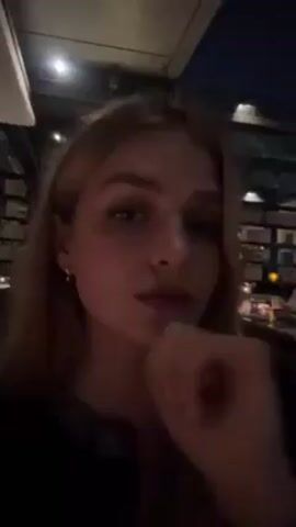 A young woman with long, light blonde hair is the main subject of the video. She is sitting at a table in what appears to be a restaurant or bar, possibly outdoors. The lighting is dim, suggesting it is evening or nighttime. She rests her face on her hand at times, looking directly at the camera. Other people are visible in the background, seated at tables and dining. The setting seems somewhat upscale, with the woman wearing what looks like a decorative top.  The overall atmosphere is relaxed and social.