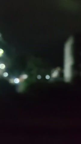 The video is an out-of-focus, nighttime recording from a moving vehicle. It shows a dark, hazy sky and a blurry, dimly lit cityscape in the distance. Several bright lights, likely from buildings or streetlights, are visible, creating streaks across the frame. The foreground is mostly dark, with indistinct shapes that might be foliage or structures. The overall impression is one of a brief, unidentifiable glimpse of an urban environment at night.