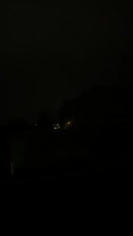 The video opens with a view of a dark, cloudy sky over a suburban landscape at dusk. A modern house with white walls and dark trim is visible on the right side of the frame, with a dark roof and windows. In the foreground, there is a dark, out-of-focus area, possibly a fence or railing, obscuring the lower portion of the scene. As the video progresses, the ambient light dims significantly, plunging the scene into near darkness. Streetlights or house lights are faintly visible in the distance, casting a dull glow. The overall atmosphere is dark and somewhat ominous due to the heavy cloud cover and fading light.