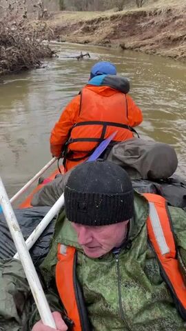 Three people are in a raft or boat, paddling down a narrow, shallow river. The riverbanks are muddy and earthen. All three people are wearing life vests. One person in the front is wearing a camouflage jacket and a black knit cap. Another person in the middle is wearing an orange jacket and a blue cap. The third person in the back is wearing a dark jacket and a black knit cap. There are dry bags and other supplies in the boat. The person in the front gives paddling directions. The sky is overcast and the surrounding vegetation is bare.