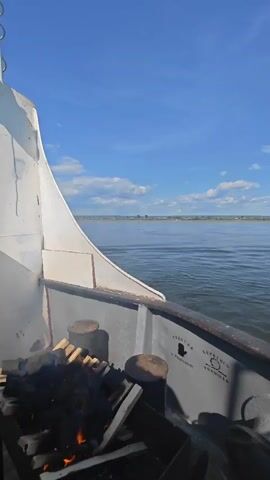 A video taken from the deck of a boat shows a tranquil scene on a wide river under a bright blue sky with scattered clouds. The foreground of the video features the railing and deck of the boat, with a metal fire pit containing burning wood and charcoal. The fire is actively burning, with flames visible. Behind the fire pit, there are several thick, cylindrical mooring posts. The boat is moving, as indicated by the slight ripple on the water's surface and the perspective shift. The middle ground of the video shows the calm river water stretching out towards the horizon. In the distance, a low, tree-lined shore is visible, with a few buildings scattered along it. The overall atmosphere is peaceful and serene.