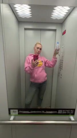 A woman with blonde hair in a bun is standing in an elevator, taking a mirror selfie. She is wearing a pink hoodie and blue jeans. She is holding a water bottle in her right hand and her phone in her left hand. The elevator has gray walls and a mirrored door. There is a panel of buttons on the right side of the elevator, and the number 4 is lit up. A advertisement for SKYNET is visible at the bottom of the mirror. The woman is looking at the camera and puckering her lips.
