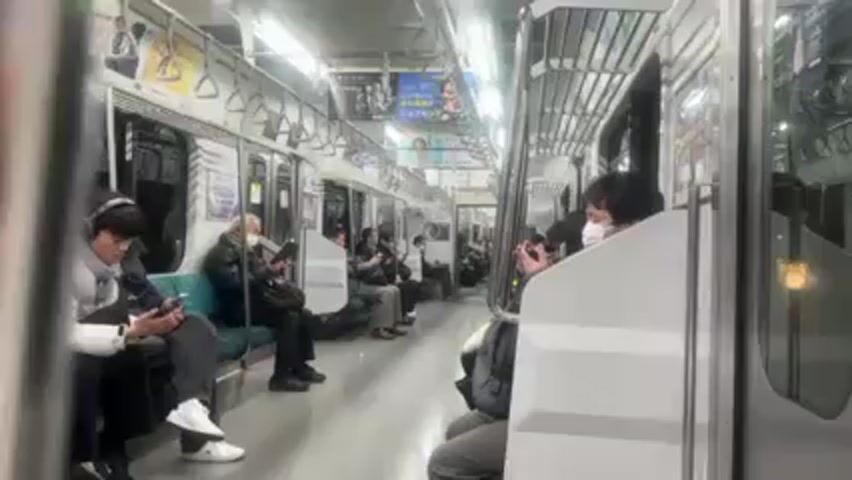 Inside a train car, passengers are seated. Some are looking at their phones or devices. Most wear masks. The train car has grey floors and light blue seating. There are advertisements on the upper walls. The train is moving.