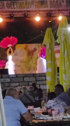 A nighttime outdoor performance is underway on a stage. Two dancers, dressed in white, are on stage. One dancer on the left is holding two large, red fans above her head. The other dancer on the right is wearing a white dress with a pink sash and holding a large, pink fan. The stage backdrop appears to be a painted scene depicting a waterfall with lush greenery. The performance is illuminated by bright stage lights. In the foreground, a group of people are seated at tables, presumably watching the show. They appear to be dining or enjoying drinks, as evidenced by bottles and glasses on the tables. The overall atmosphere seems festive and relaxed, with the audience engaged in the entertainment. The sky above is dark, indicating it is evening or night. The audience members are casually dressed, suggesting an informal event. There are large, yellow umbrellas with white stripes in the right side of the frame, possibly providing shade or shelter, though it's nighttime. A banner with text is visible at the top of the stage, but it is partially obscured and difficult to read clearly, though it appears to be in Cyrillic. The setting suggests a public gathering or festival in Gara Oreshets, Bulgaria.
