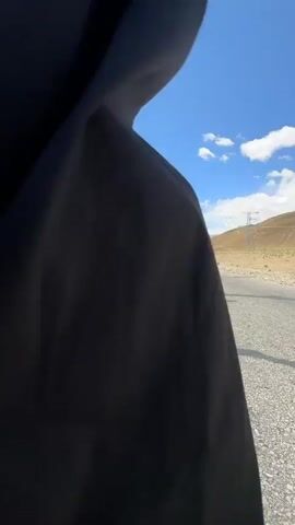 The video begins with a close-up of a dark blue fabric. It then cuts to a wide shot of a person, appearing to be a woman with long hair, walking away from the camera on a straight asphalt road. She wears a vibrant, multi-colored long-sleeved top and loose black pants. Her hair is blowing in the wind, and her shadow is prominent on the road, which features a distinct yellow line. The setting is an expansive, arid landscape with distant mountains under a clear blue sky dotted with white clouds. Power lines or towers are visible in the far background. The person stops, turns to the side, and then strikes a joyful pose, balancing on one leg with arms outstretched, smiling at the camera. The bright sunlight indicates it is daytime, and the overall scene conveys a sense of freedom and open space in a remote, mountainous region of Mainpu township, China.