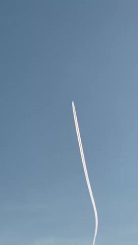 A thin white contrail curves upwards against a clear blue sky. The contrail appears to be from an aircraft. The bottom of the contrail is out of frame.