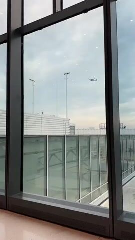 The video shows a view through a large window at an airport. Outside, two airplanes are visible in the sky. One airplane is higher and further away, while the other is lower and closer. There are tall light poles and a ribbed white structure, likely part of the airport's terminal building, in the foreground. The sky is overcast with clouds. The interior of the airport, visible through the window, includes glass walkways and dark metal framing. The lighting suggests it is daytime.