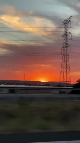 This video captures a sunset viewed from a moving vehicle, likely a car. The sun, a bright orange orb, is just touching the horizon, casting a vibrant orange and pink glow across the sky. Wispy clouds are scattered throughout, tinged with the sunset colors. A large metal electricity pylon is visible in the foreground on the left, and another smaller one on the right. Power lines stretch across the frame. A treeline and some distant structures are visible on the horizon. The foreground shows a blurred road or highway and some vegetation, indicating the vehicle's motion. The overall impression is a dynamic and colorful sunset scene observed during travel.