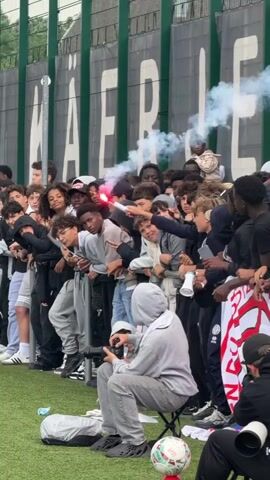 A short vertical video shows a crowd of young individuals gathered by a fence next to a green field. The crowd stands behind a metal barrier, leaning over it. The background features a large green fence with white text that appears to say "KAERLE". The ground is green turf. One person in the crowd holds a red flare that emits red light and white smoke. The smoke rises into the air. A person in a gray hoodie sits on a folding chair in the foreground, facing away from the camera, seemingly filming the scene with a camera. Another person, partially visible, sits next to the camera person, wearing a black top and a white turban. A football with a red and white pattern is visible on the ground near the seated individuals. The crowd consists mainly of teenagers and young adults, with a diverse mix of genders and ethnicities. Some individuals in the crowd are smiling and looking at the camera or the flare. The overall atmosphere seems lively and celebratory.