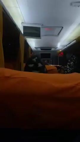 The interior of a bus is visible. It appears to be a sleeper bus, with orange cushions in place of seats. Two individuals can be seen: one in the foreground, partially obscured, and another further down the aisle wearing a patterned shirt and looking down. There are monitors affixed to the ceiling. The bus interior is dimly lit.