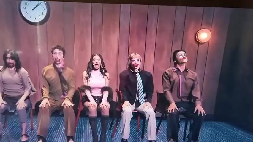 The video features five individuals – four men and one woman – seated on chairs and performing synchronized, energetic movements on what appears to be an indoor stage or set. The backdrop consists of vertical wooden panels, with a round analog clock displaying approximately 10:10 visible on the left wall, and a decorative wall light or object on the right.

The central figure is a woman with long dark hair, wearing a pink sleeveless top and black shorts or a short skirt. She frequently displays a wide-eyed, open-mouthed, and somewhat theatrical expression. The four men, two positioned on either side of her, are dressed in varying casual to semi-formal attire, including shirts, and some with ties.

The performance involves highly synchronized, jerky, and almost robotic movements, characterized by rapid head swings, upper body contortions, and arm gestures. Their faces are often blurred due to the speed of their actions, contributing to a somewhat distorted or puppet-like aesthetic. The overall activity suggests a choreographed dance or performance piece.

The lighting is theatrical, brightly illuminating the performers against the wooden backdrop. The atmosphere is energetic and performative, carrying an element of the bizarre due to the precise and slightly unsettling nature of the synchronized movements.

The location context for this media is Xàtiva, Spain. No other legible text is visible in the frame apart from the clock face.