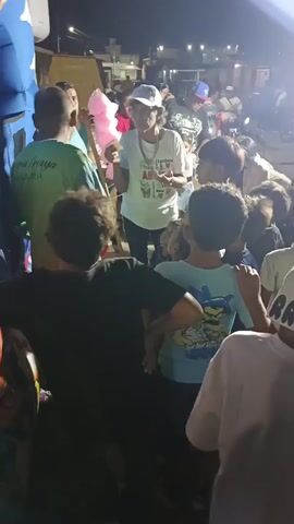 At night, a crowd of people surrounds a woman who appears to be distributing candy, with a cotton candy dispenser in the background. Many of the people are children, and the woman seems to be interacting with them. In the immediate foreground is a large blue and white inflatable bounce house. The overall environment seems to be outdoors, possibly at a street fair or community event.