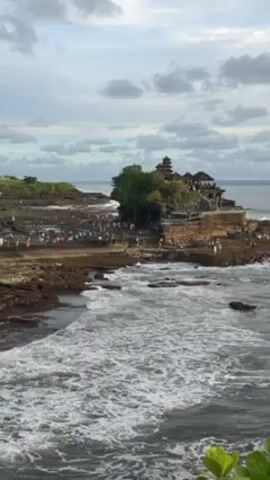 The video shows a wide shot of the Tanah Lot temple in Bali, Indonesia, perched on a rocky outcrop surrounded by the ocean. Many people are present on the rocky base surrounding the temple. The ocean has moderate waves, and the sky is overcast with a mix of gray and white clouds.