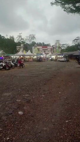 The video captures an outdoor scene, likely a fairground or market, under a cloudy sky. In the foreground, the ground is covered with gravel and dirt, with some scattered debris. Towards the middle ground, there are several stalls or booths set up, some with tents and awnings, illuminated by strings of lights. A Ferris wheel can be seen in the background, along with other amusement park-like structures. Several motorcycles are parked on the left side of the frame, and a few people are visible walking around or near the stalls. The overall atmosphere appears to be that of a community gathering or event. The lighting suggests it might be late afternoon or early evening.