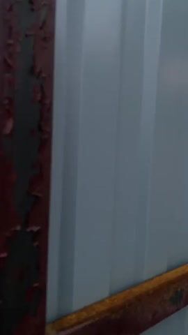 This is a video clip from Uchaly, Russia, capturing a rural setting during what appears to be sunset. The initial frames show a close-up of weathered, rusty metal structures, possibly part of a fence or building, with peeling red paint. The background is a light-colored corrugated metal wall.

The scene then transitions to an outdoor view, focusing on a dirt path winding through lush green vegetation. In the distance, several buildings, possibly houses or outbuildings, are nestled among trees. One building has a distinctive arched greenhouse structure next to it. The sky is the most striking element, transitioning from a soft, hazy daylight to a vibrant sunset. The clouds are painted with intense shades of orange, red, and pink, creating a dramatic and colorful display. The light suggests it's late evening. The overall mood evoked is one of rustic beauty and the peacefulness of a rural sunset. There is no visible text in the video.