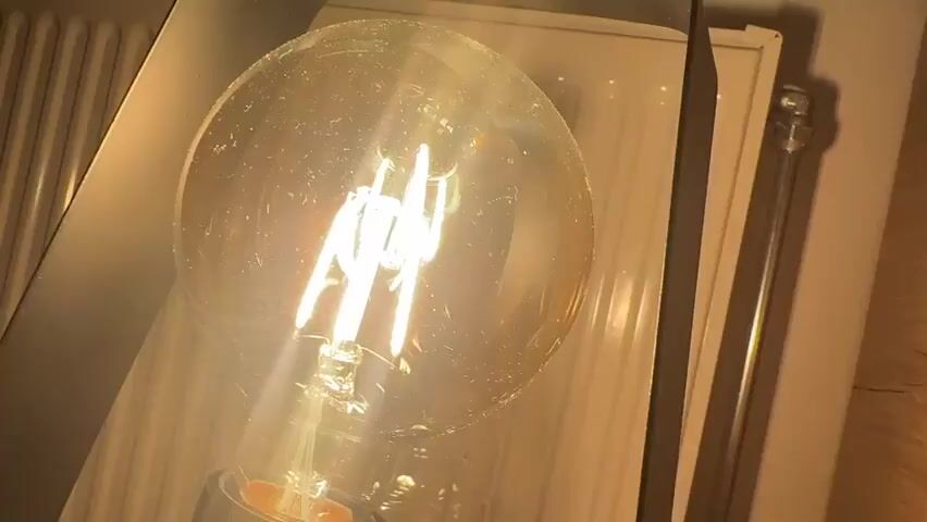 A close-up shot of a lit Edison-style light bulb. The bulb has a clear glass housing and a visible filament. The filament is glowing brightly, emitting a warm yellow light. The bulb is positioned in front of a white radiator, which has vertical fins. Part of a metallic fixture or lampshade is visible to the left of the bulb. The background is slightly out of focus, showing the radiator and what appears to be a wooden surface. The overall impression is one of a warm, indoor environment.