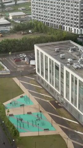 The video captures an aerial view of an outdoor area. In the foreground, there are two rectangular green sports courts with various exercise equipment, including pull-up bars and benches. A paved walkway with a geometric pattern connects these courts to a white building with numerous windows. To the left of the courts, a grassy area is visible. In the background, there is a parking lot with several cars, some trees, and construction vehicles. Further in the distance, a tall, modern residential building stands out. The overall scene suggests an urban or suburban environment with recreational facilities.