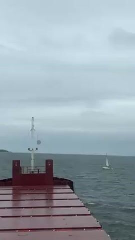 The video shows a view from the deck of a large cargo ship on a gray, overcast day. The sea is choppy. A small sailboat with white sails is visible in the distance to the right. The ship's deck is red and slightly wet. There is a small, boxy structure with a white antenna on top of it in the center of the view. In the far distance, there is a blurry landmass.
