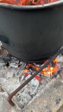 A large pot of bright red crawfish is shown cooking outdoors over a wood fire. The crawfish are packed tightly in the pot, and steam rises from them, indicating they are boiling. The pot is made of dark metal and is supported by a metal rack over the flames. The fire is visible beneath the pot, with glowing embers and active flames. The ground around the fire appears to be rough and stony. In the background, out of focus, are several people, some standing and some seated, and what appears to be a large green object, possibly a watermelon. The overall scene suggests an outdoor cooking event, likely a crawfish boil.
