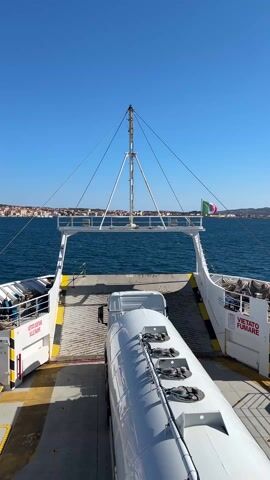 The video shows the view from the deck of a ferry, facing towards the stern and a distant coastline. A white tanker truck is parked on the deck, facing away from the camera, towards the open ramp at the stern. The ramp is flanked by white structures with diagonal yellow and black hazard stripes. A sign reading "VIETATO SOSTARE SULLE RAMPE" is visible on one side, and "VIETATO FUMARE" on the other, indicating no standing on the ramp and no smoking. Above the ramp, a white mast with rigging extends upwards. An Italian flag with a white shield featuring a red cross waves gently from the top of the mast. The sea is a deep blue, and the sky is clear and bright. In the background, a town is visible on the coastline.