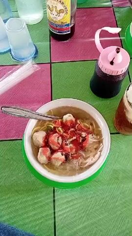 This is a top-down video of a table setting. The table is covered with a checkered tablecloth in shades of pink and green. On the table are several bowls of food, drinks, and condiments. In the foreground, a bowl of noodles with meatballs and red sauce is visible, with a spoon resting in it. To the right of this bowl is a glass filled with an iced beverage. Further back on the table, there are bottles of liquid, a pink dispenser with a dark liquid, and another bowl of noodles. A person is sitting on the floor, cross-legged, eating noodles from a bowl with a fork and spoon. The person is wearing a blue t-shirt and dark pants. The environment appears to be outdoors or in an open-air structure, as indicated by the natural lighting and background elements. The video is shot from a stationary, high angle, offering a clear view of the table and the meal.