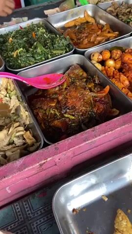 The video shows a close-up view of several metal trays filled with various food dishes. The trays are arranged on a surface, possibly a table or a market stall. In the foreground, a pink serving utensil is partially submerged in a dark, saucy dish containing what appears to be meat or fish and some vegetables. To the left and right of this dish, other trays contain different food items, including leafy green vegetables, a dish with small round objects that resemble quail eggs in a sauce, and a dish with what looks like fried items. In the background, more trays of food are visible, as well as parts of people's hands and arms, suggesting that this is a food stall or a buffet. The lighting is somewhat dim, and the focus is primarily on the food. There is a hint of a colorful patterned surface beneath the trays. The video appears to be a brief, shaky clip, possibly captured by someone browsing or selecting food.