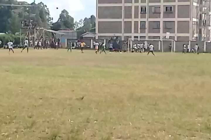 The video shows a soccer game being played on a grassy field with a building in the background. People in different colored jerseys are running around the field, kicking a ball. There are spectators along the sidelines. The weather appears to be overcast.