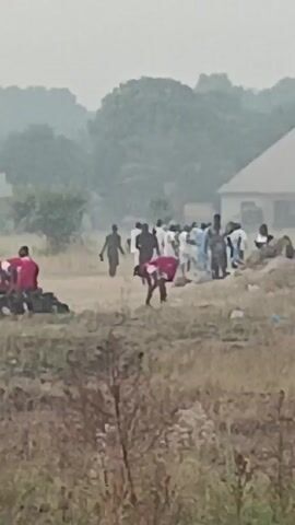 A group of people are gathered in a field on a hazy day. Some are standing, while others are bent over or sitting. A few people are wearing red shirts, while the majority appear to be in white or light-colored clothing. There are bags or objects piled on the ground near the people in red shirts. In the background, trees and a building with a pitched roof are visible. The foreground is filled with dry, overgrown vegetation.