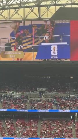 The video shows a large stadium filled with spectators. A giant screen displays a group of women in matching outfits, holding pom-poms and performing a routine. The screen also shows "HYDRATION BREAK" and a timer counting down from 1:40. The women are on a raised platform, and the overall atmosphere appears to be energetic and festive, likely during a sporting event.