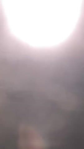 A blurry, out-of-focus video shows a bright light source, possibly the sun, in the upper left quadrant of the frame. The rest of the frame is filled with hazy, indistinct shapes and light flares. A small, geometric object, appearing to be a sticker or label, is visible in the upper center of the frame, reflecting the bright light. The background is obscured, but appears to be an outdoor scene with some horizontal lines suggesting land or a horizon. The video is shaky, indicating movement.