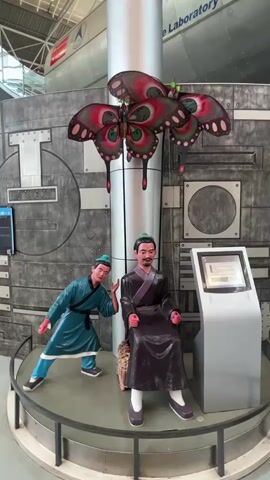 The video shows an indoor exhibit featuring two life-size mannequins and a large decorative butterfly sculpture. One mannequin, dressed in a dark purple robe and a green headscarf, is seated on a basket-like chair. The other mannequin, wearing a teal-colored robe and a green headscarf, is positioned to the left, leaning forward with its right arm extended, as if gesturing. Both mannequins have a reddish hue on their faces. Above the mannequins, three large, colorful butterfly sculptures with prominent red, green, and black patterns hang from a central pillar. The background consists of a textured gray wall, possibly part of a museum display, with circular and rectangular metallic elements. In the upper background, a large, silver cylindrical object, possibly an aircraft or rocket component, is visible with the words "Laboratory" inscribed on it. The mannequins and the butterfly sculpture are displayed on a circular platform with a dark railing. A digital display screen is positioned to the right of the seated mannequin. The overall lighting suggests an indoor exhibition space with overhead illumination.