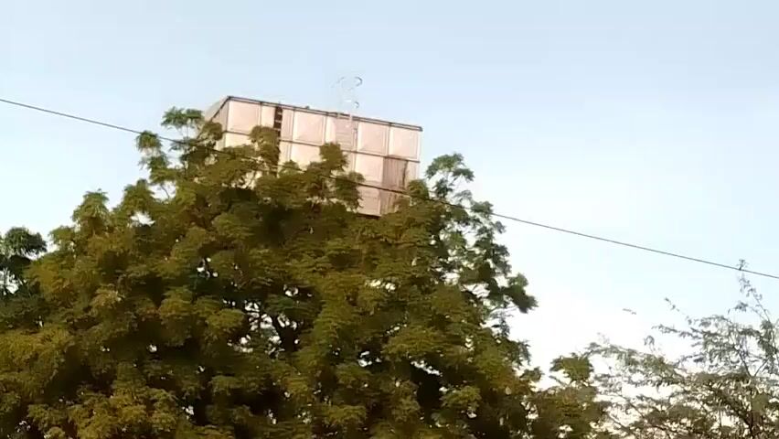The video shows a large, cubic kite stuck in the branches of a green leafy tree. The kite is made of a light-colored material, possibly paper or plastic, and appears to be constructed from multiple panels. A thin wire runs across the top of the frame, possibly a power line, and extends behind the kite and tree. The background is a clear, pale blue sky, suggesting it's daytime. The camera is positioned below the kite and tree, looking upwards. There are no people visible in the frame.