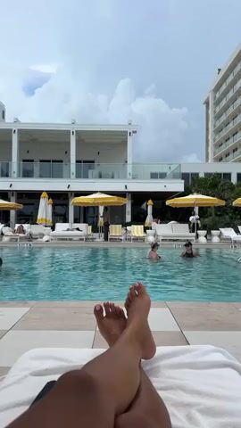 A first-person perspective video shows a person's feet crossed on a towel at the edge of a swimming pool. The pool is filled with clear blue water, and several people are swimming in it. Behind the pool, there is a resort or hotel with white buildings, yellow umbrellas, and lounge chairs. The sky is partly cloudy.